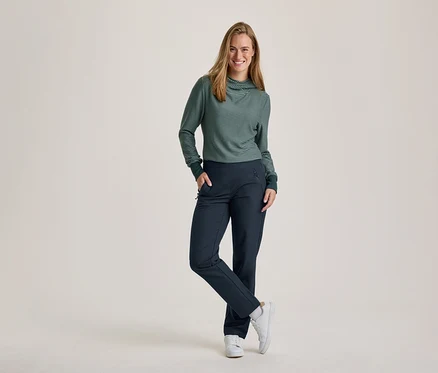 Une femme souriante aux cheveux longs se tient debout, une main dans la poche et une jambe croisée. Elle porte un haut à manches longues rayé vert et blanc avec un col roulé, un pantalon bleu foncé et des baskets blanches.