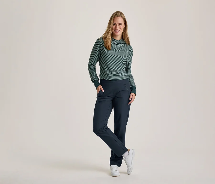 Une femme souriante aux cheveux longs se tient debout, une main dans la poche et une jambe croisée. Elle porte un haut à manches longues rayé vert et blanc avec un col roulé, un pantalon bleu foncé et des baskets blanches.