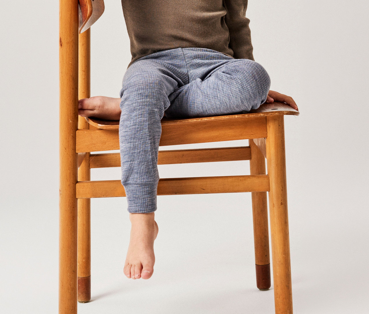 Enfant assis sur une chaise en bois, portant un haut marron à manches longues et un pantalon bleu à motifs, un pied pend.