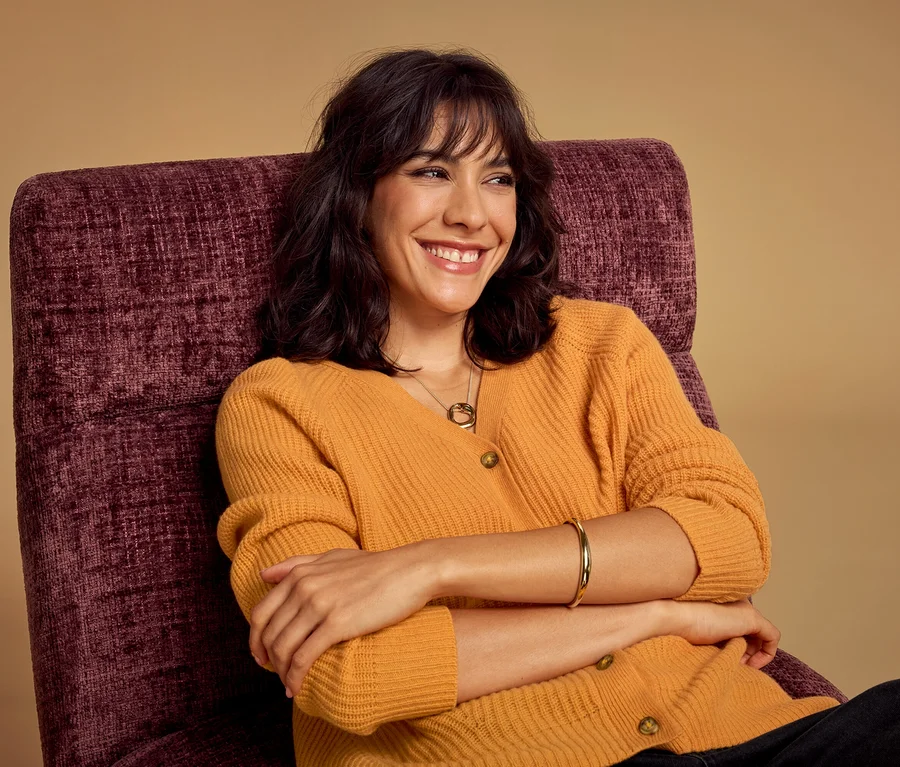 Une femme souriante aux cheveux foncés est assise dans un fauteuil violet, portant un cardigan jaune moutarde et un collier.