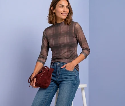 Une femme souriante est assise sur un tabouret, portant un T-shirt en maille Street One à carreaux, un jean bleu et tenant un sac seau bordeaux.