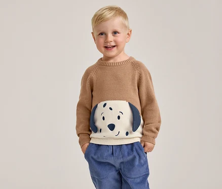 Un jeune garçon souriant aux cheveux blonds se tient les mains dans les poches, portant un pull marron avec un motif de visage de chien blanc et bleu sur le devant et un pantalon en velours côtelé bleu.