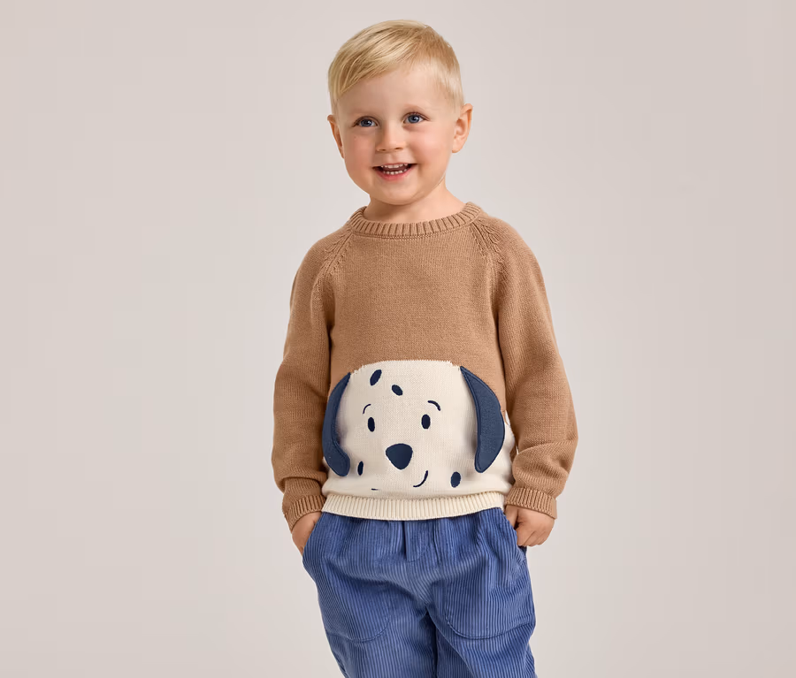 Un jeune garçon souriant aux cheveux blonds se tient les mains dans les poches, portant un pull marron avec un motif de visage de chien blanc et bleu sur le devant et un pantalon en velours côtelé bleu.