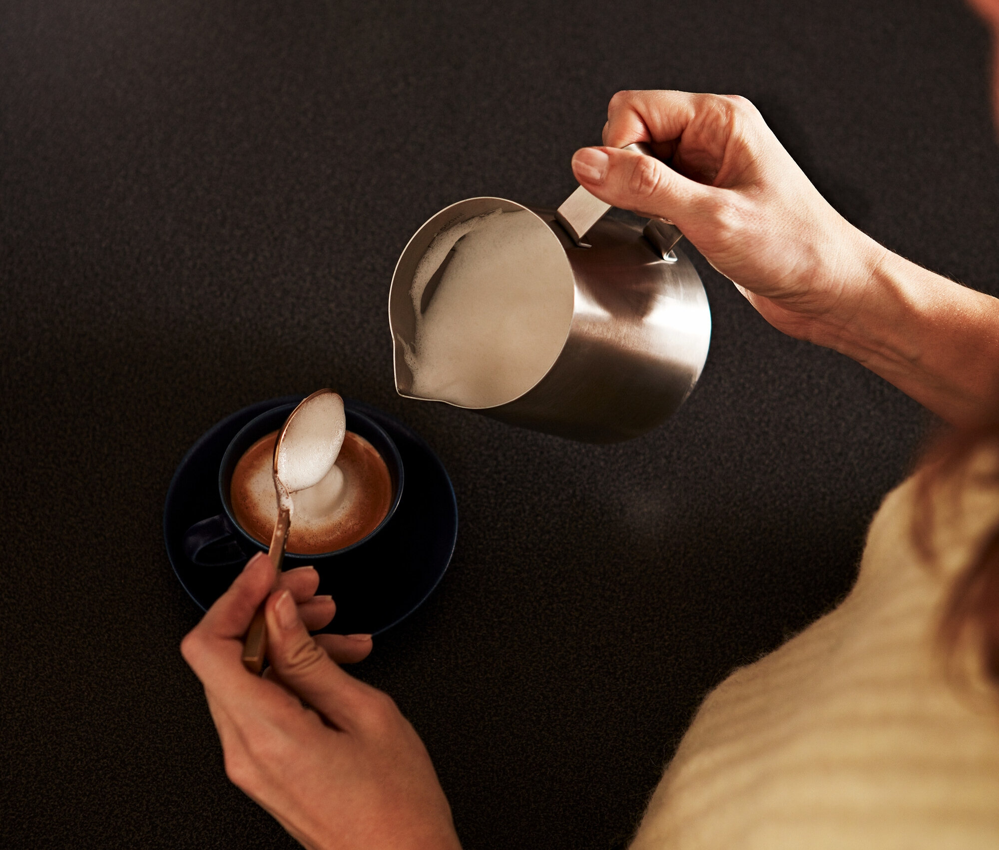 Une femme verse du lait mousseux d'un petit pichet à lait en acier inoxydable dans une tasse de café et ramasse de la mousse avec une cuillère.