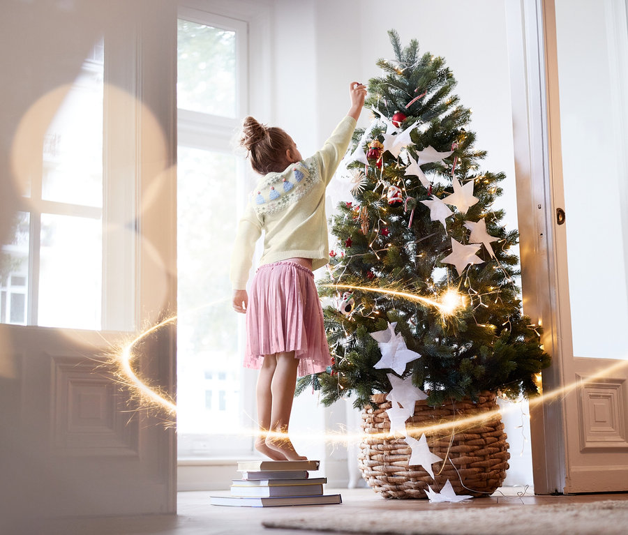 Une fille debout sur des livres décore un sapin de Noël. Elle porte une jupe en tulle et un pull en maille.