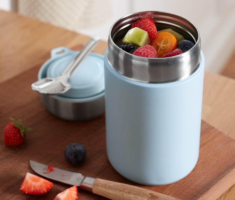 Un récipient thermo à emporter ouvert rempli de fruits posé sur une planche à découper en bois à côté d'un couvercle et d'une cuillère.