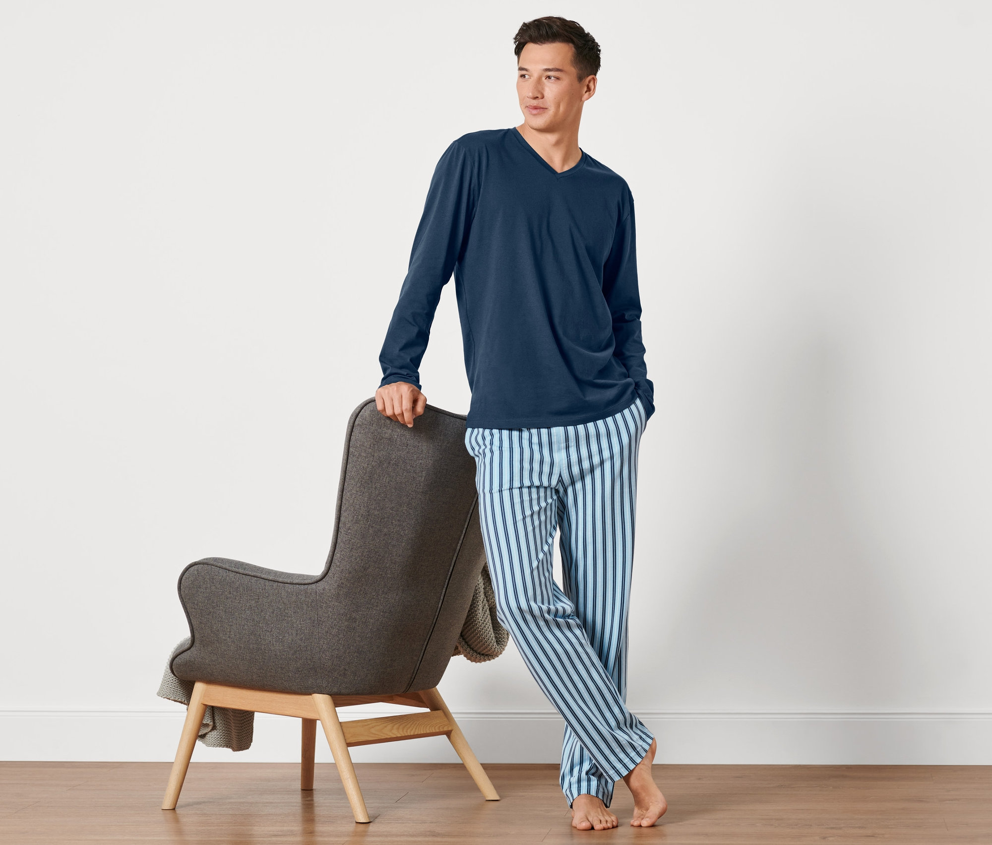 Un homme pose en pyjama à côté d'un fauteuil gris.