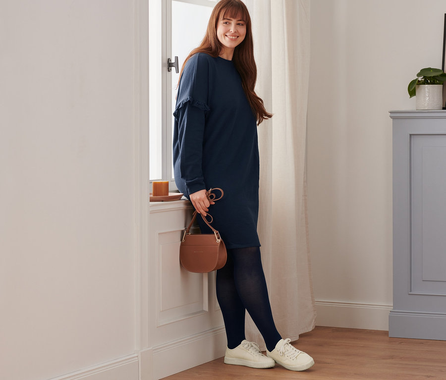 Une femme souriante est appuyée contre une fenêtre. Elle porte une robe molletonnée, des baskets en cuir, un collant opaque (lot de 2) et un sac à bandoulière.