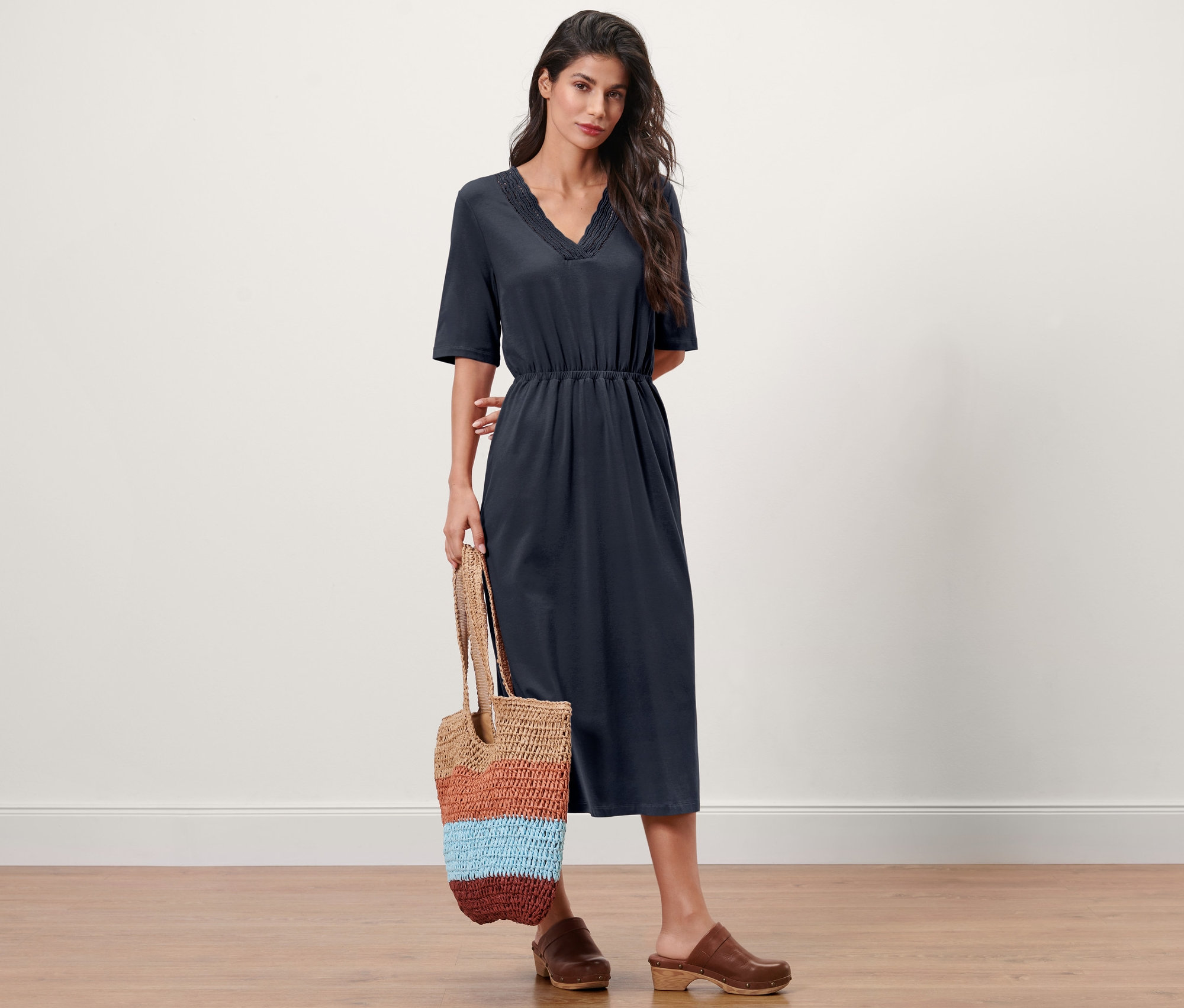 Femme posant en robe en jersey bleu marine et sabots marron tenant un sac rayé.