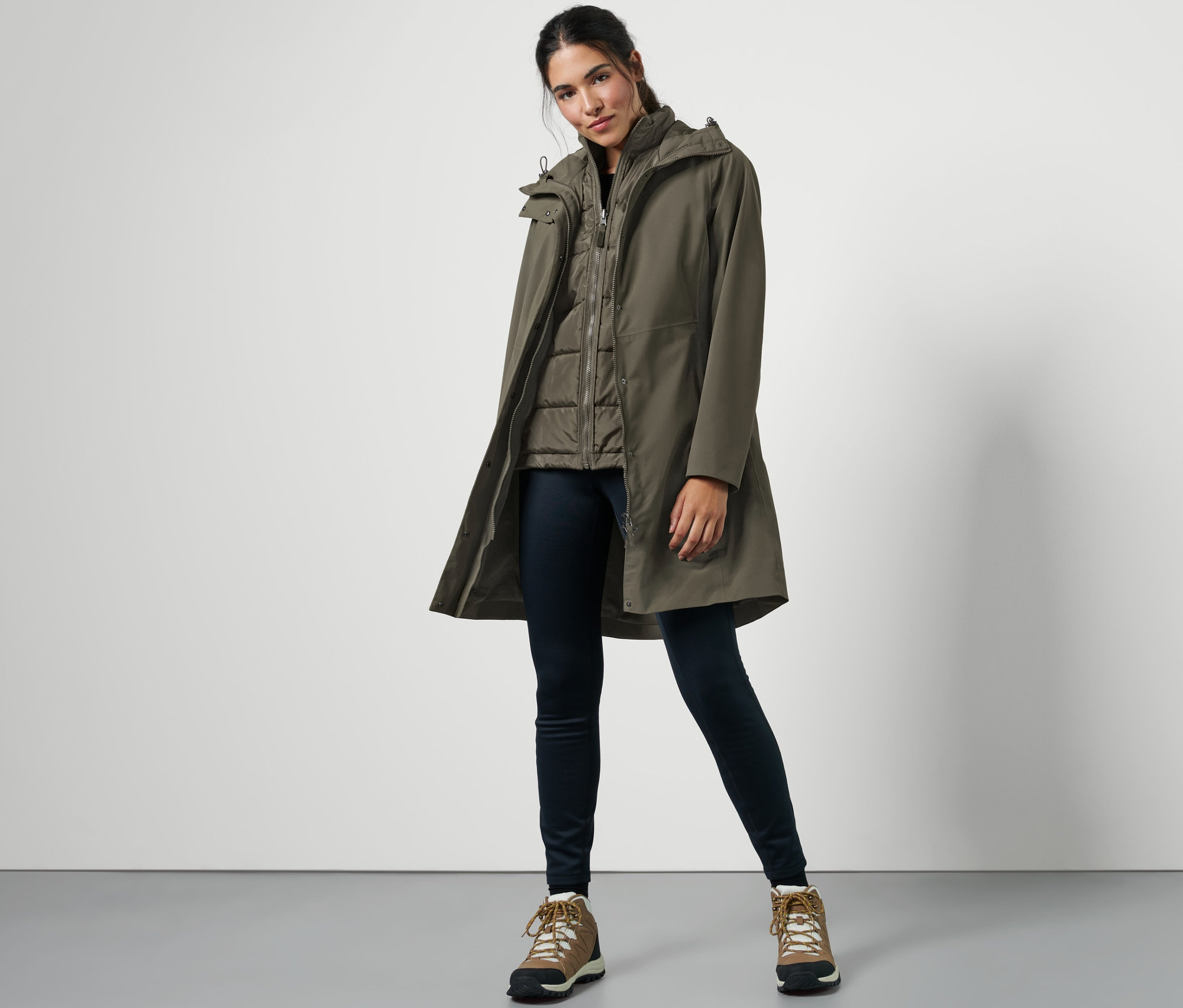 Une femme pose portant un manteau de pluie 3-en-1 avec gilet, un jean foncé et des chaussures de randonnée.