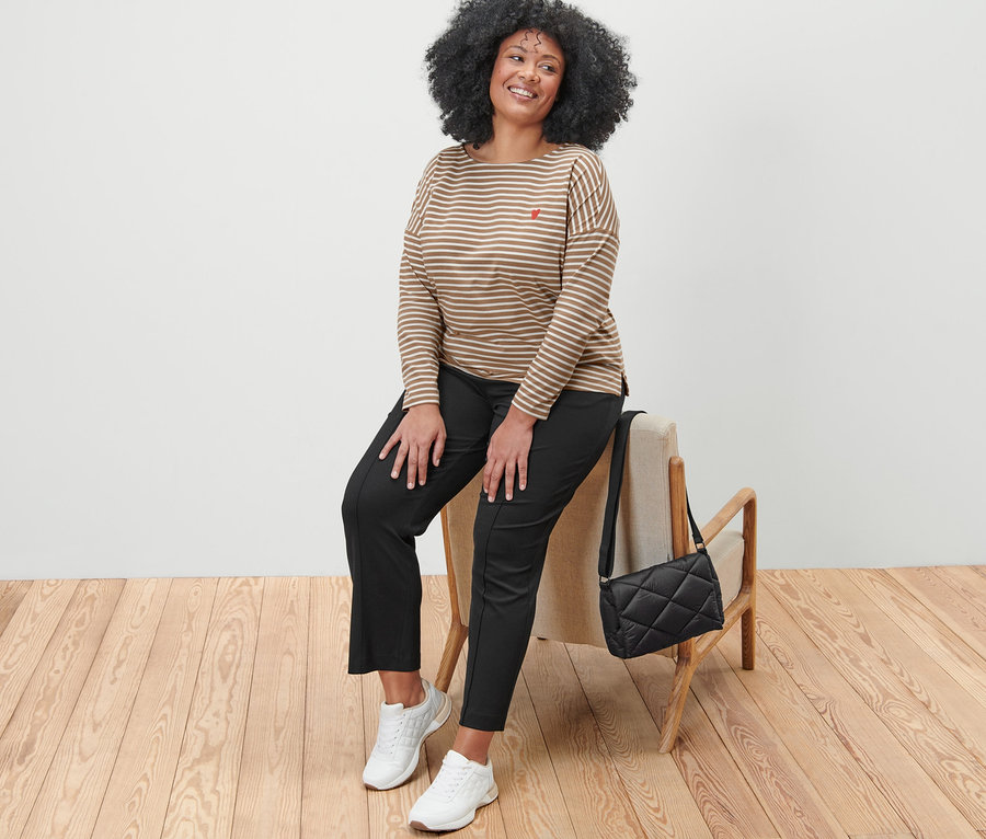 Une femme est assise sur une chaise et porte un T-shirt à manches longues rayé, un pantalon de jogging noir et des baskets blanches. À côté d'elle se trouve un sac matelassé à bandoulière.
