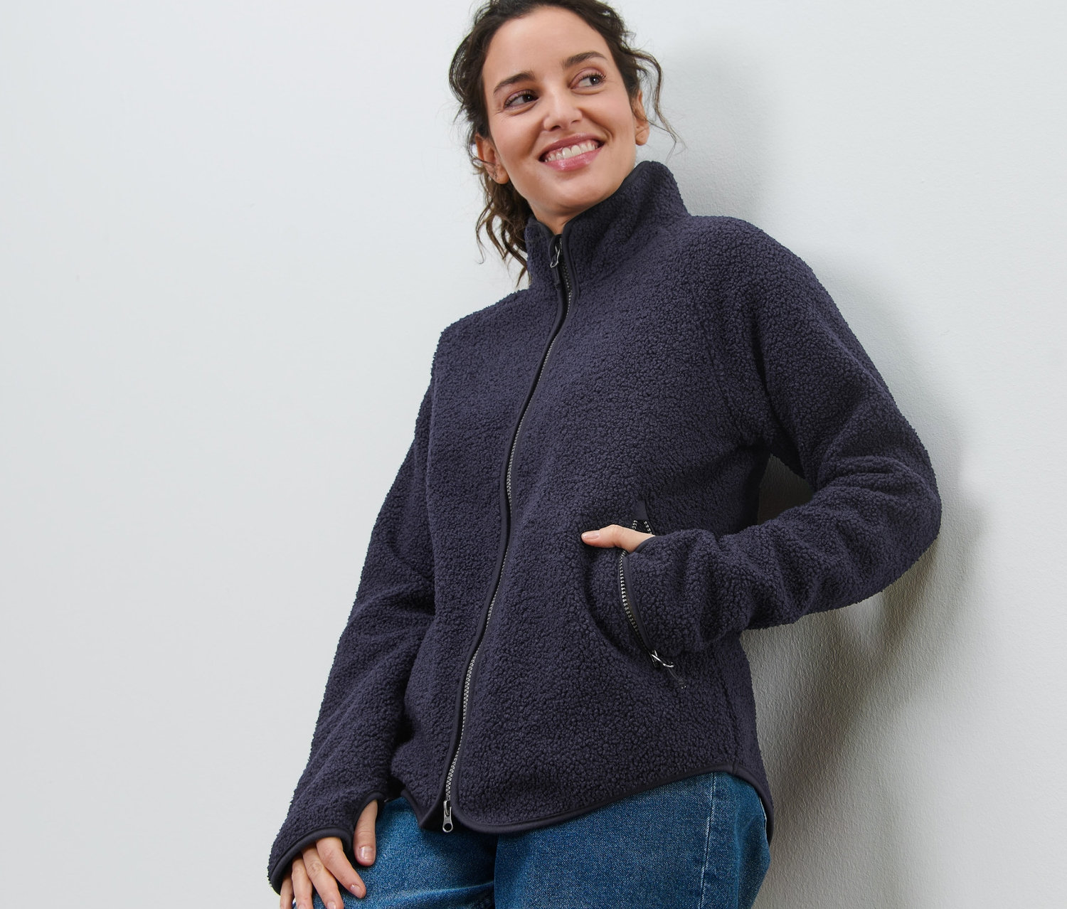 Une femme souriante appuyée contre un mur porte une veste en polaire teddy bleu marine et un jean.