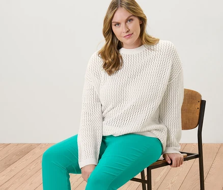 Une femme est assise sur une chaise et porte un pull en maille pointelle et un pantalon stretch 7/8 turquoise.