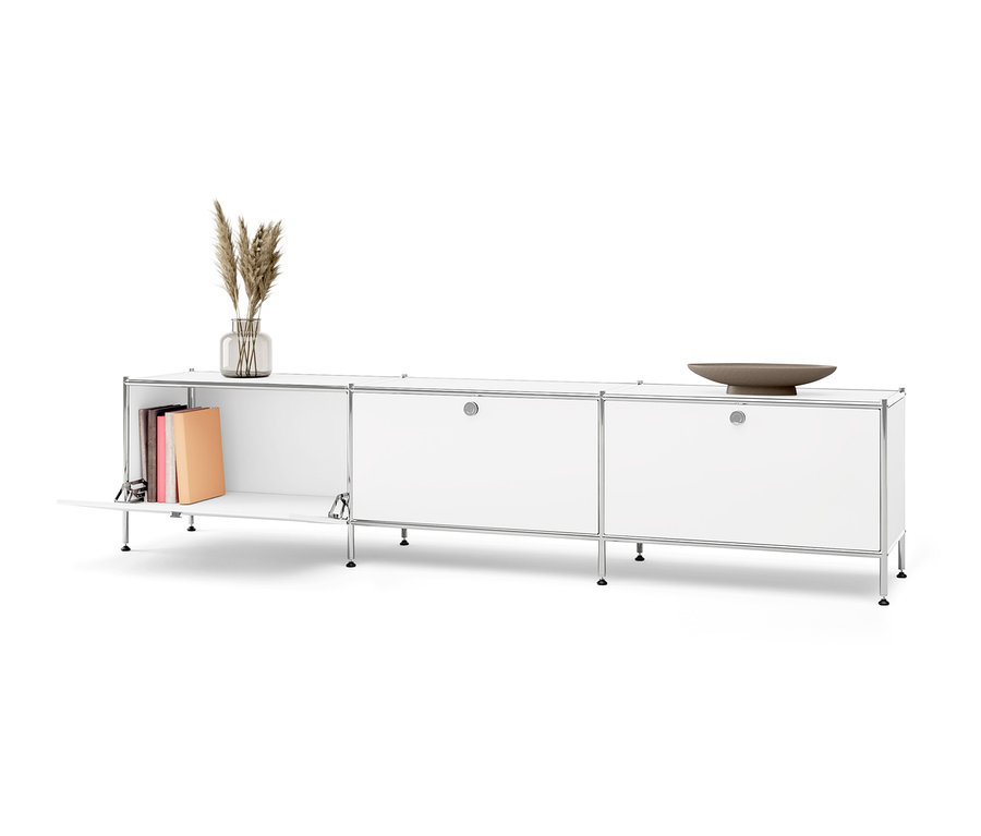Meuble bas en métal blanc « CN3 » à 3 abattants, dont un ouvert avec des livres à l'intérieur. Un vase avec de l'herbe et un bol marron sont posés sur le meuble.