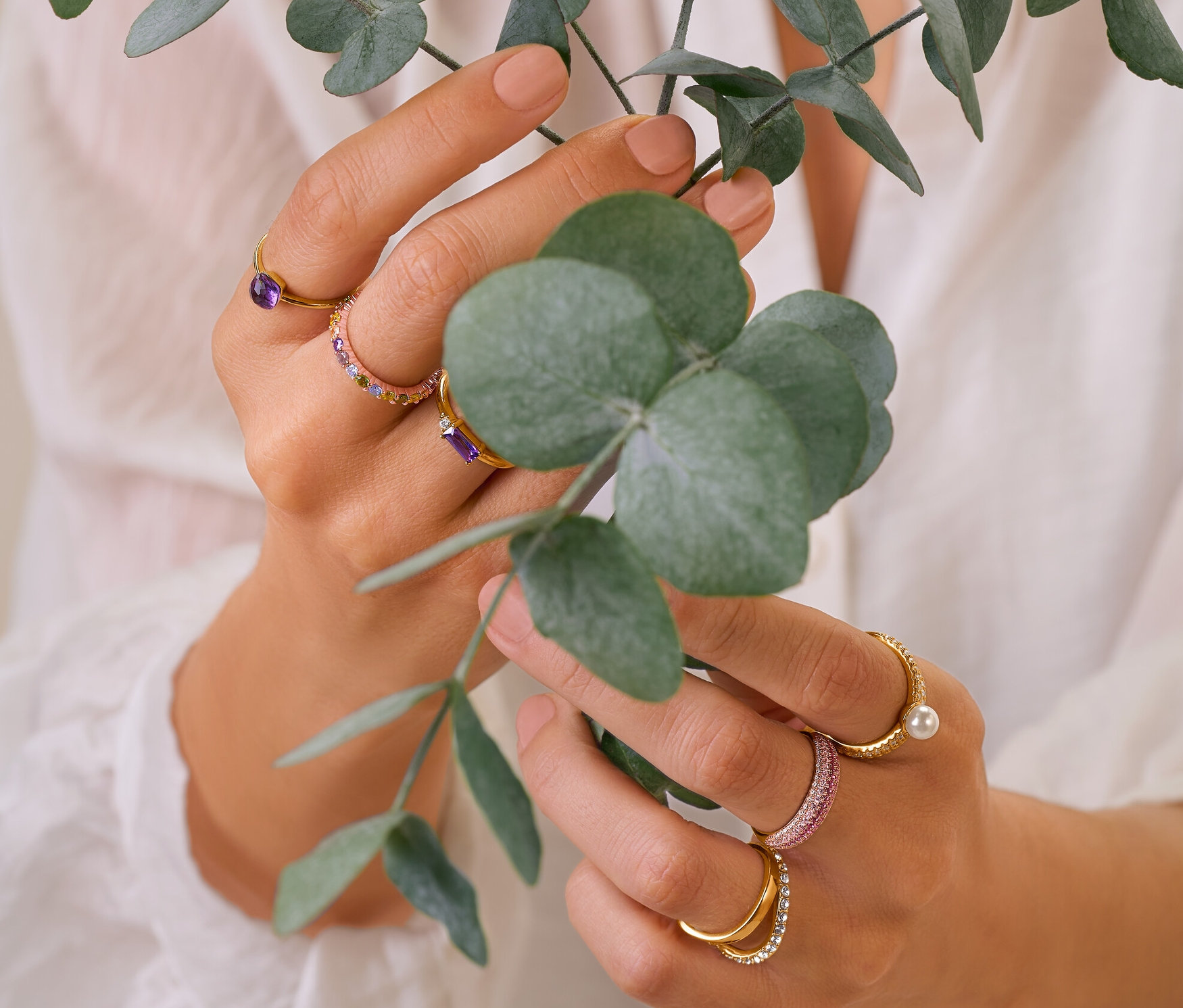 Une femme tient des branches d'eucalyptus et porte des bagues en argent 925, une bague multicolore, une bague double vague avec cristaux et une bague en or 375 et améthyste et une bague en or 585 et améthyste.