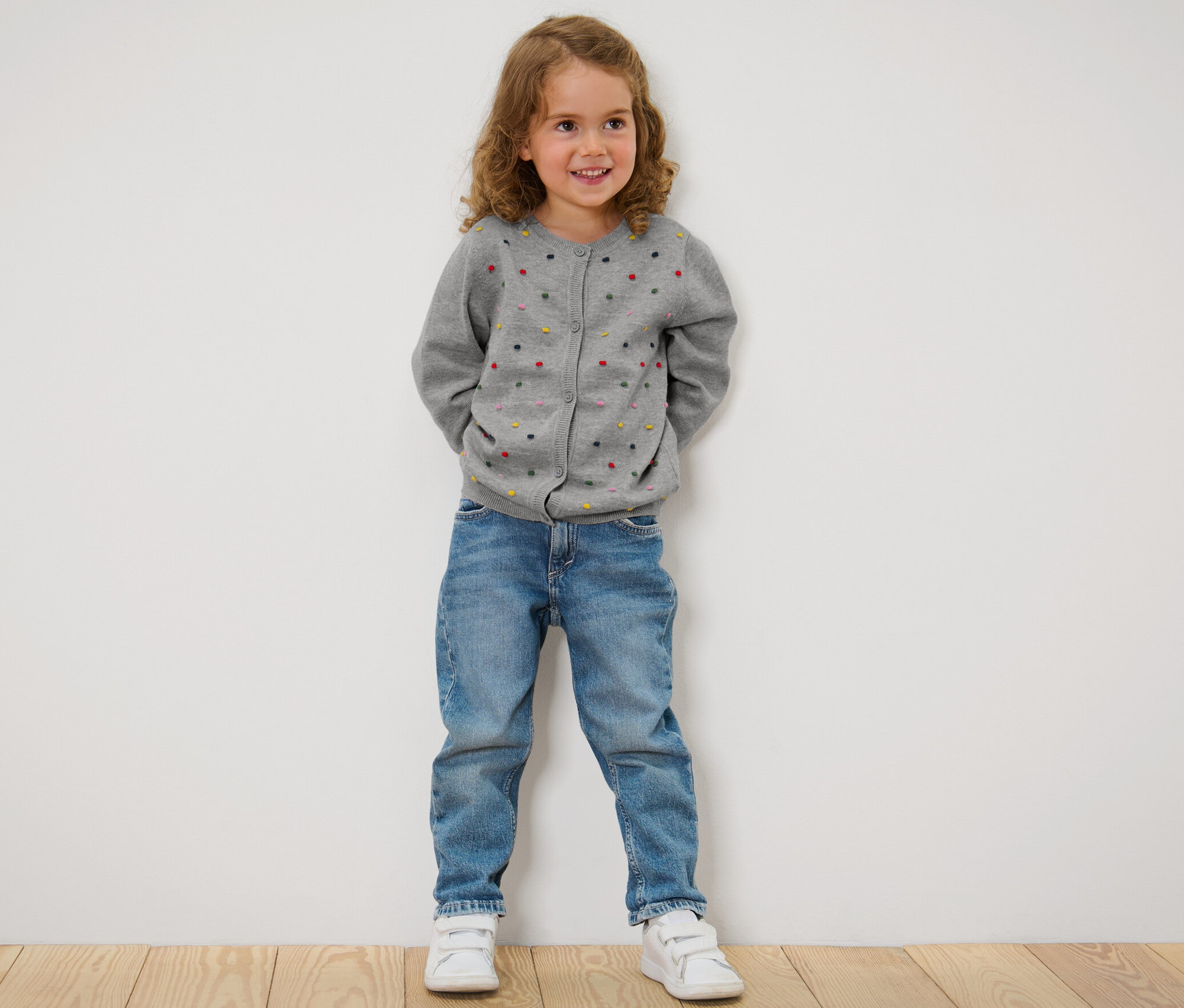 Fillette debout, les mains derrière le dos, portant une jaquette en maille pour enfant, un jean et des baskets.