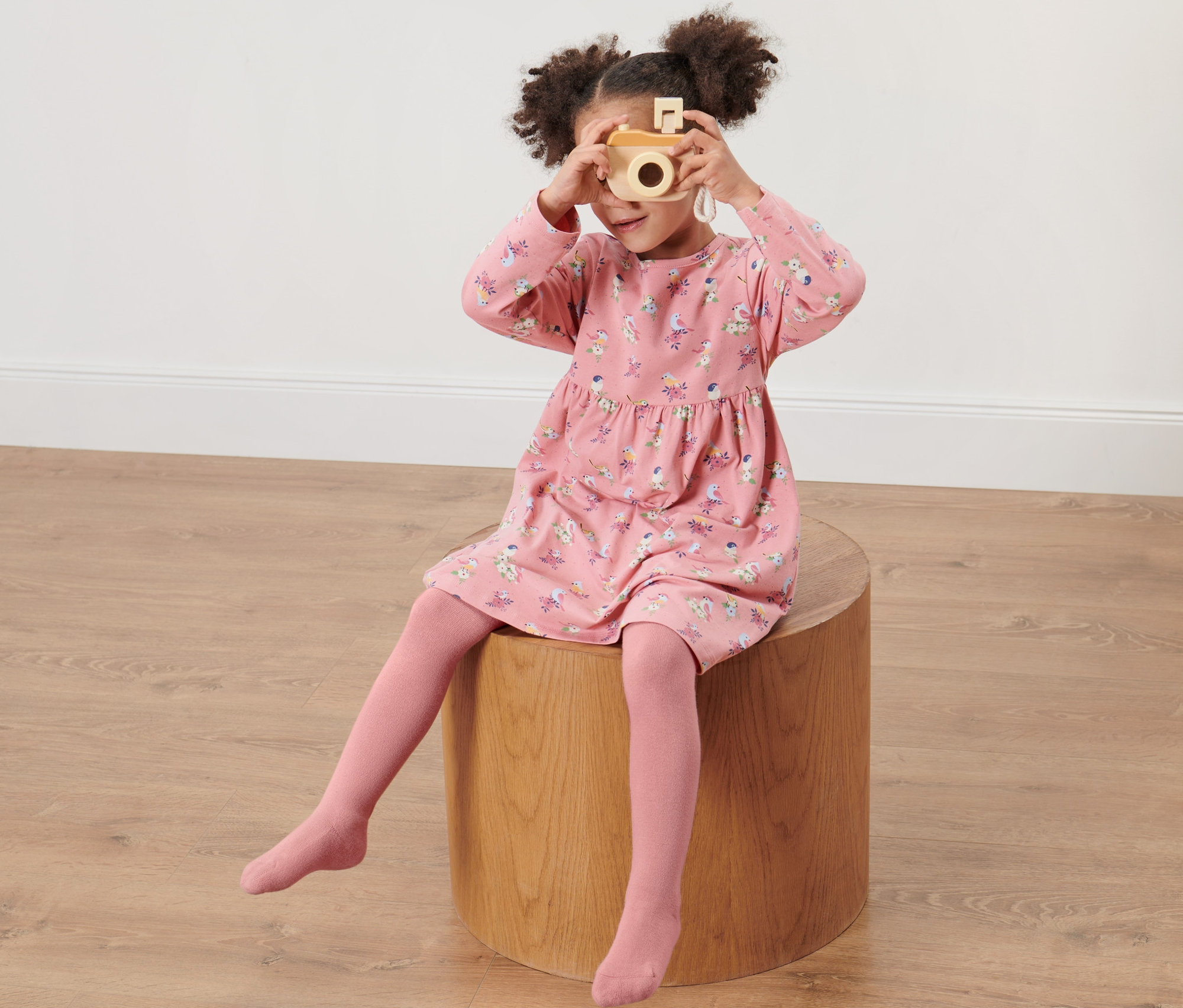 Une fille est assise sur un tabouret en bois et prend une photo avec un appareil photo en bois. Elle porte une robe rose imprimée et 2 collants pour enfant, rose/bleu clair.