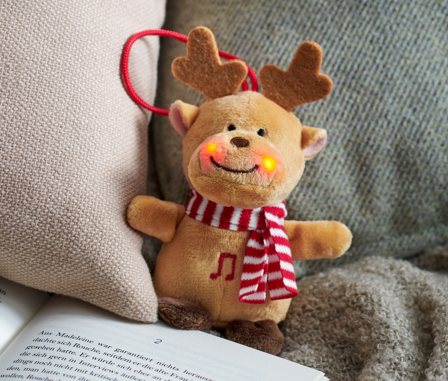 Mini peluche chantante renne posée sur un livre ouvert à côté d'un coussin beige.