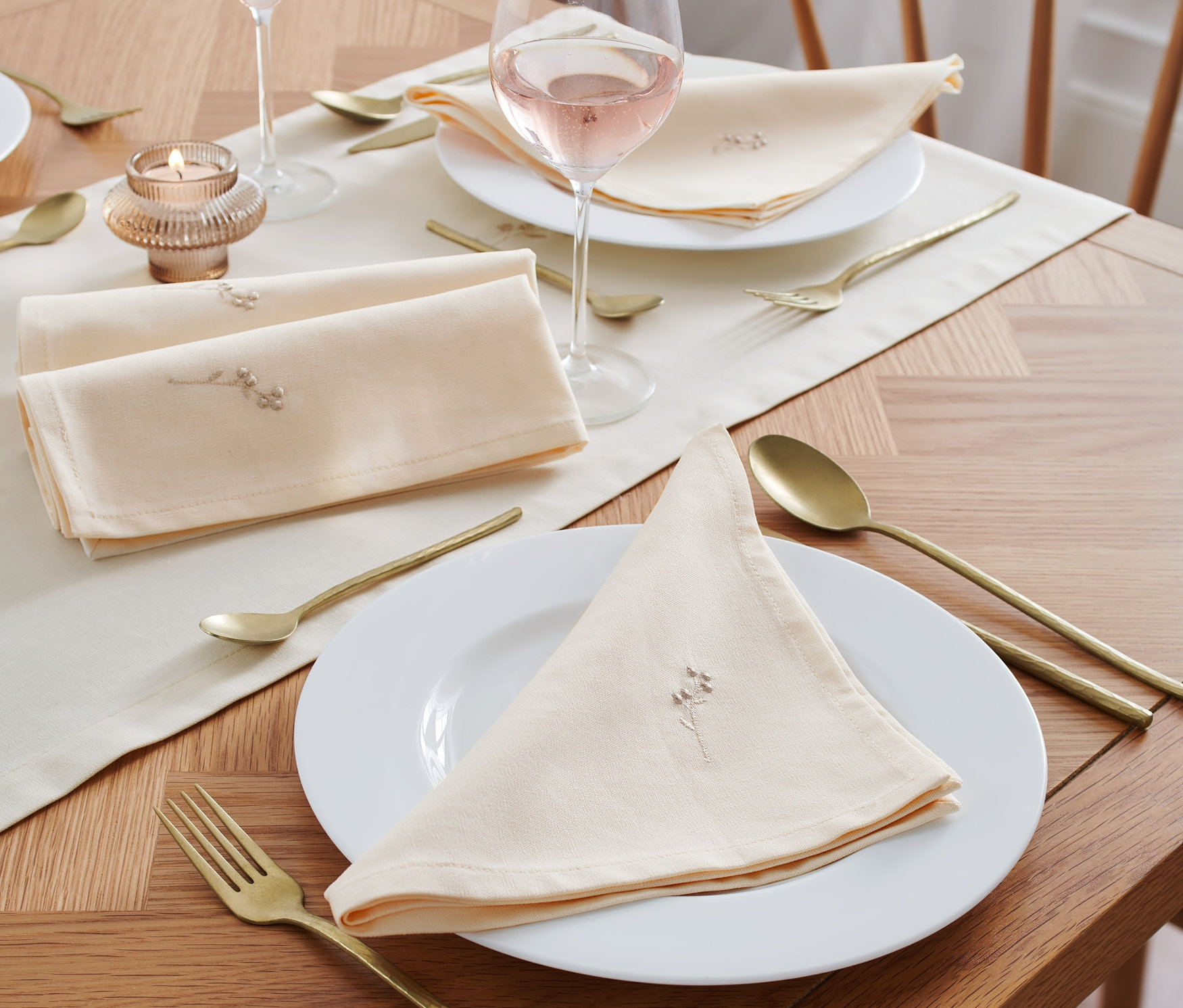 Sur une table sont disposées 4 serviettes brodées, des verres de vin rosé, des couverts dorés et une bougie.