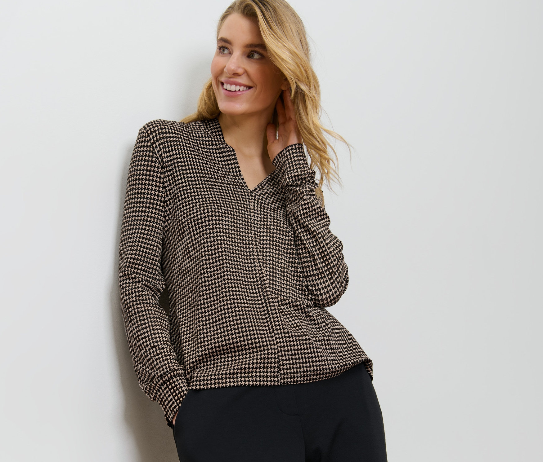 Une femme blonde, appuyée contre un mur, porte une blouse imprimée avec encolure en V.