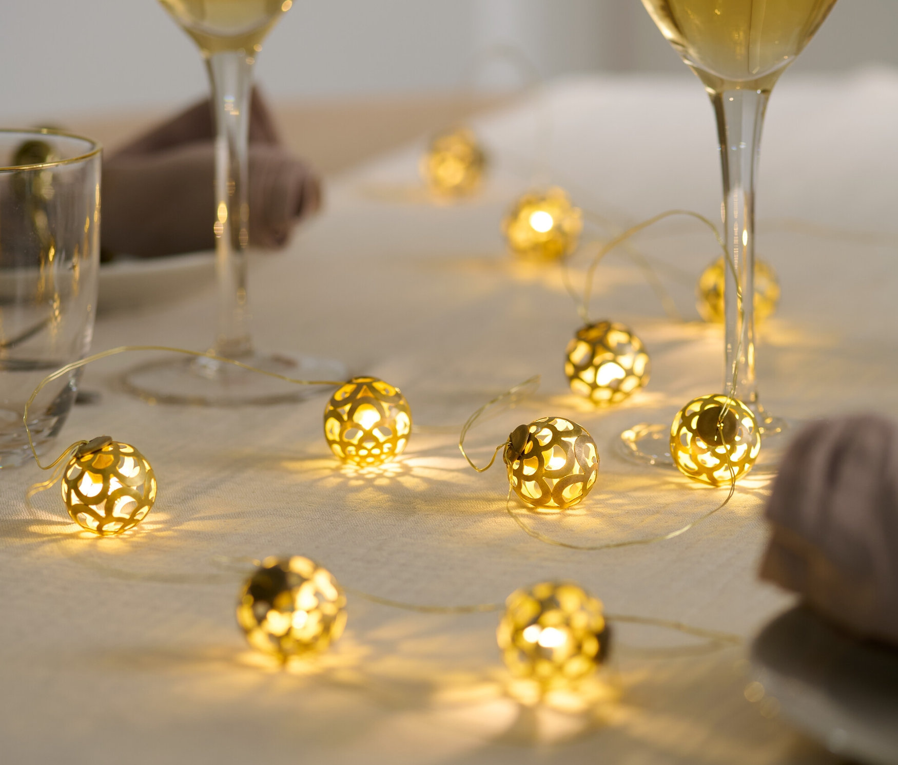 Sur une table sont posées une guirlande lumineuse à LED, deux verres de vin blanc et des serviettes.