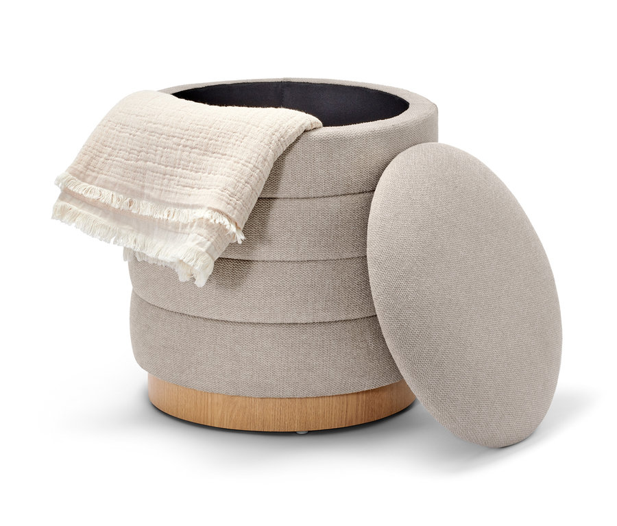 Un pouf beige avec espace de rangement et une base en bois. Une couverture blanche repose sur le pouf et le couvercle est à côté.