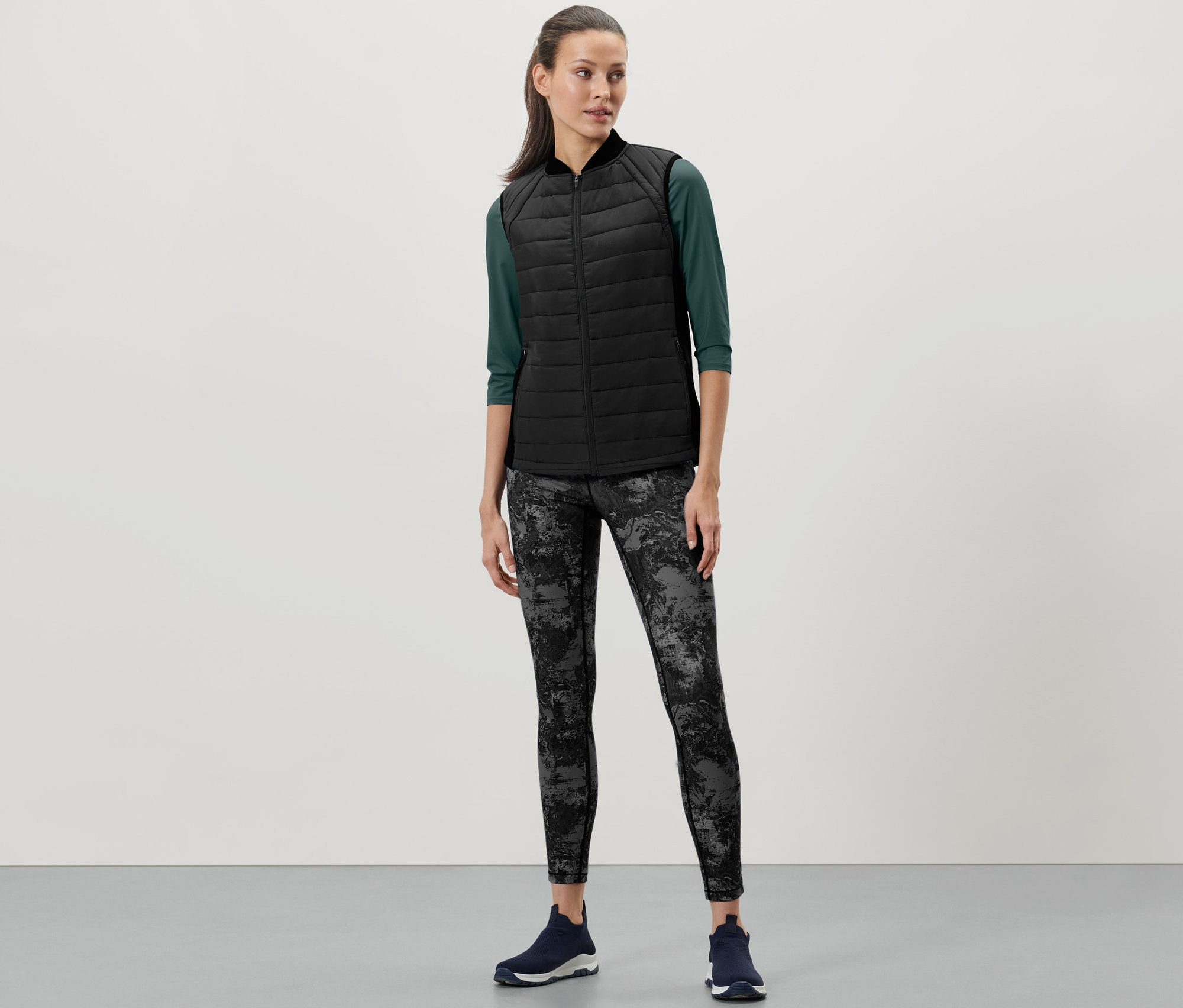 Une femme porte un gilet sans manches matelassé thermique, un t-shirt vert à manches trois quarts, un legging à motifs et des baskets bleu marine.