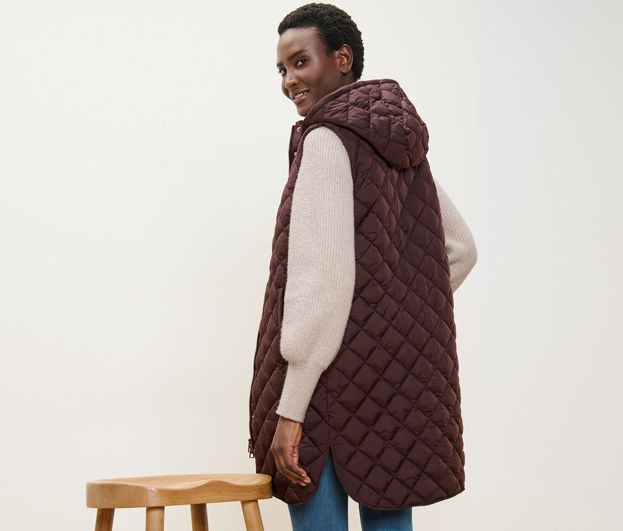 Femme souriant par-dessus son épaule et portant un long gilet sans manches matelassé avec capuche.