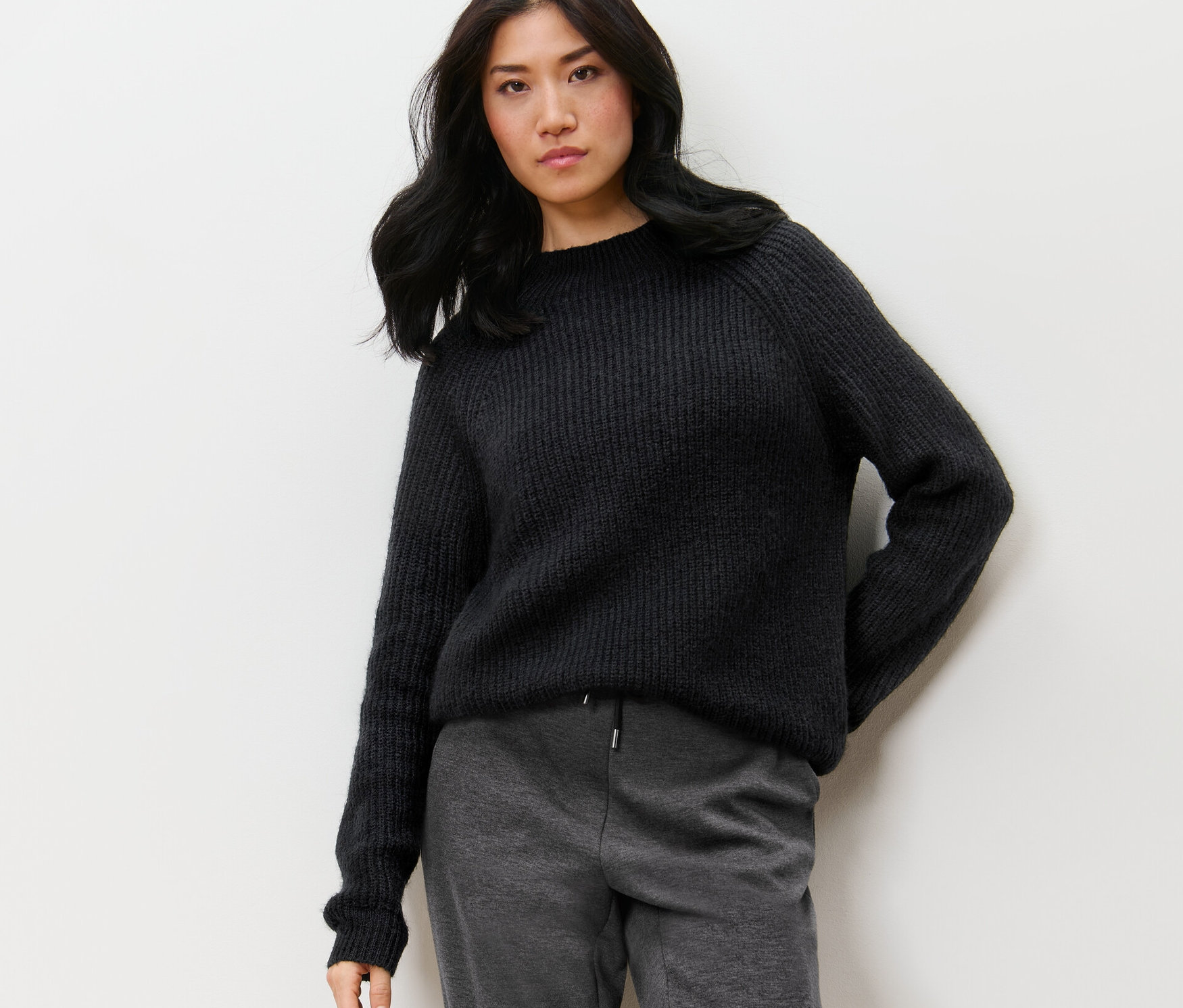 Une femme pose devant un mur et porte un pull noir à col droit en grosse maille.