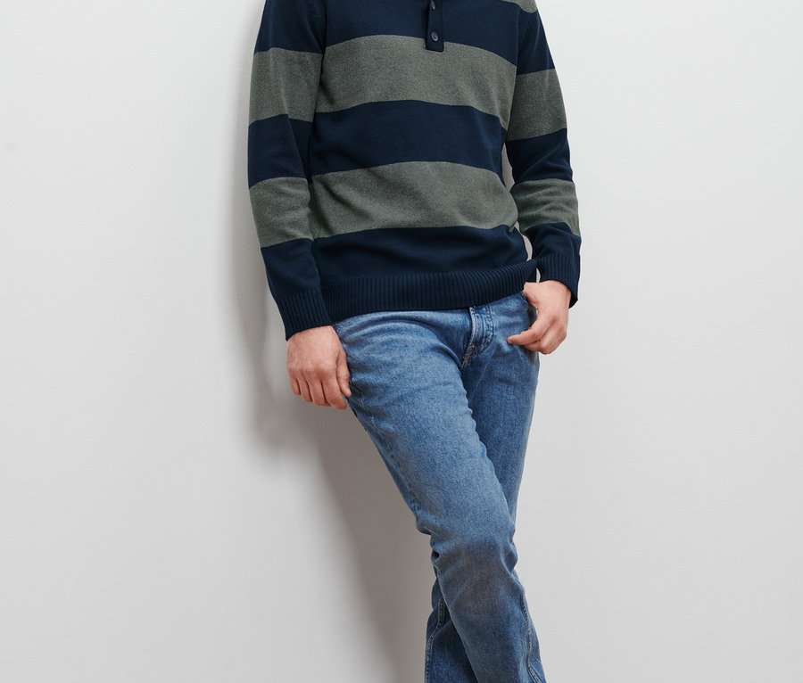 Un homme appuyé contre un mur porte un pull rayé et un jean.