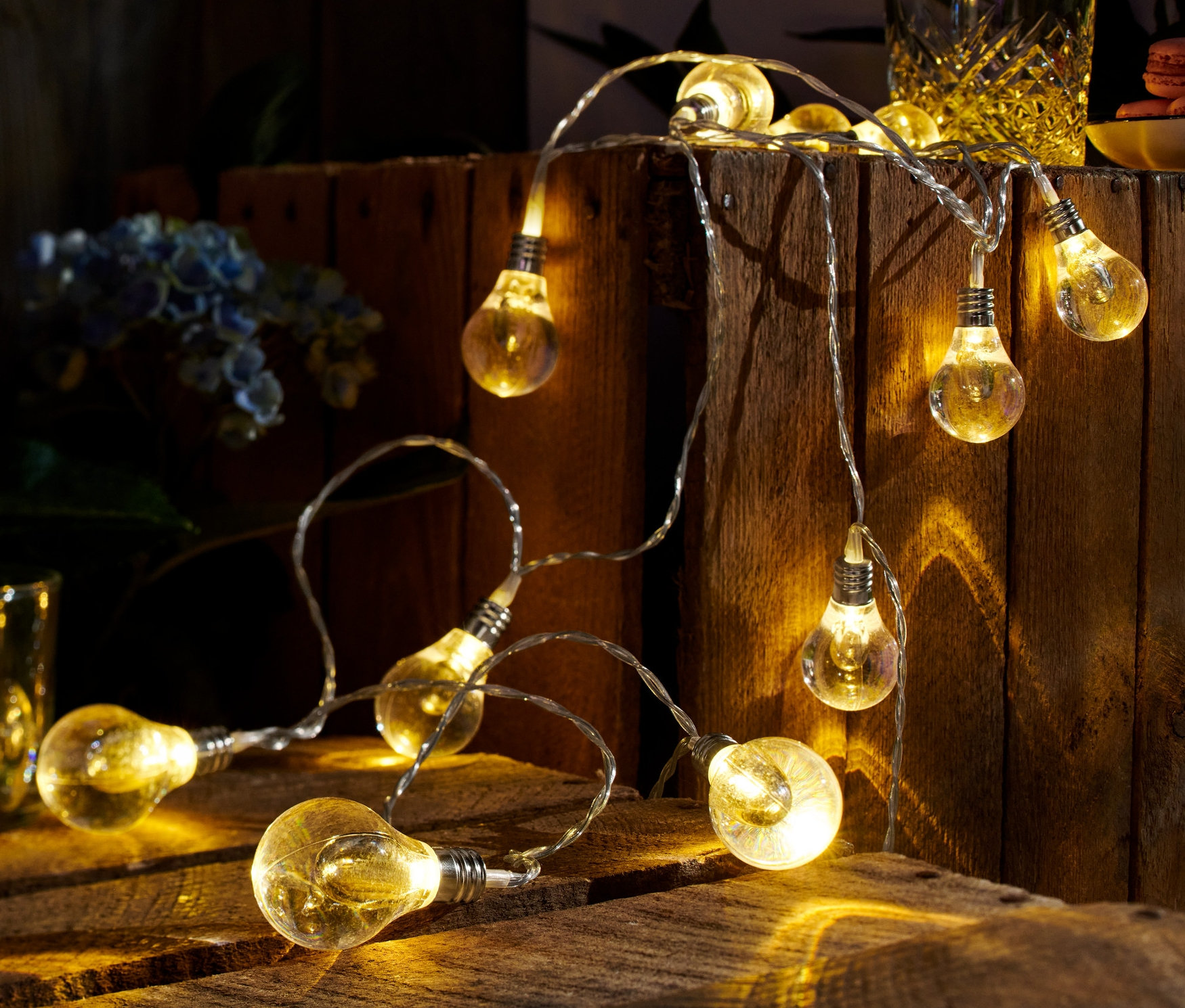 Une guirlande lumineuse à LED est posée sur une table en bois, avec un verre de boisson et des fleurs en arrière-plan.