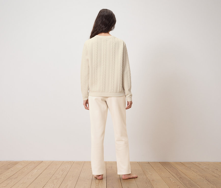 Une femme se tient dos à la caméra, portant un pull et un pantalon couleur crème.