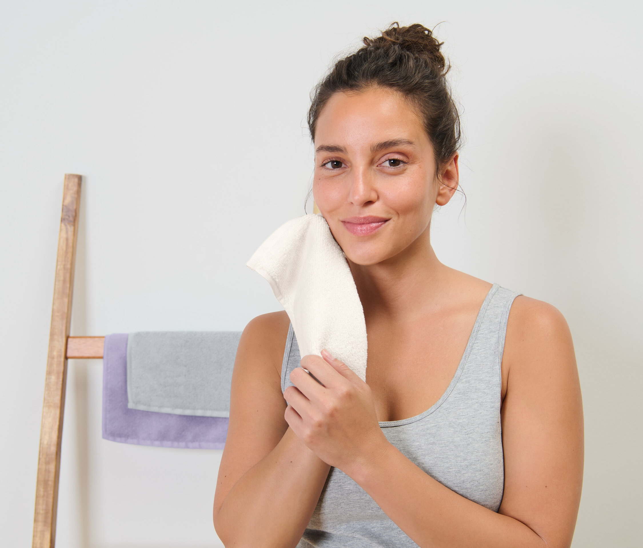 Une femme s'essuie le visage avec une serviette blanche. Derrière elle se trouve un porte-serviettes en bois avec une serviette grise et une serviette violette.