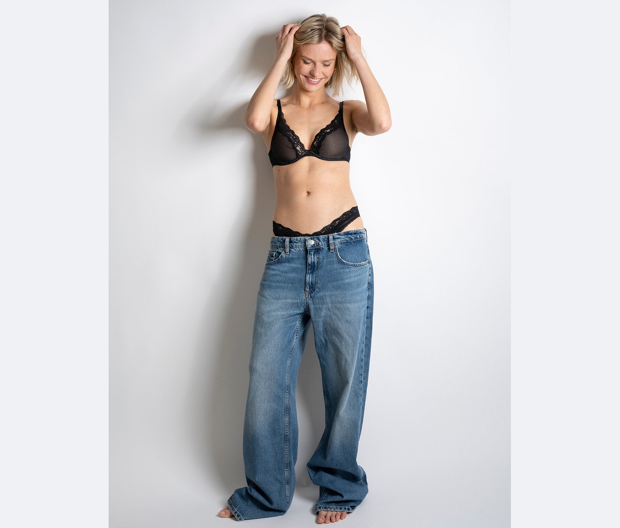 Une femme sourit et tient ses mains sur sa tête, elle porte un soutien-gorge et un string en dentelle noire et un jean ample.