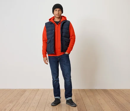 Un homme portant un sweat à capuche orange, un gilet matelassé bleu marine et un jean bleu.