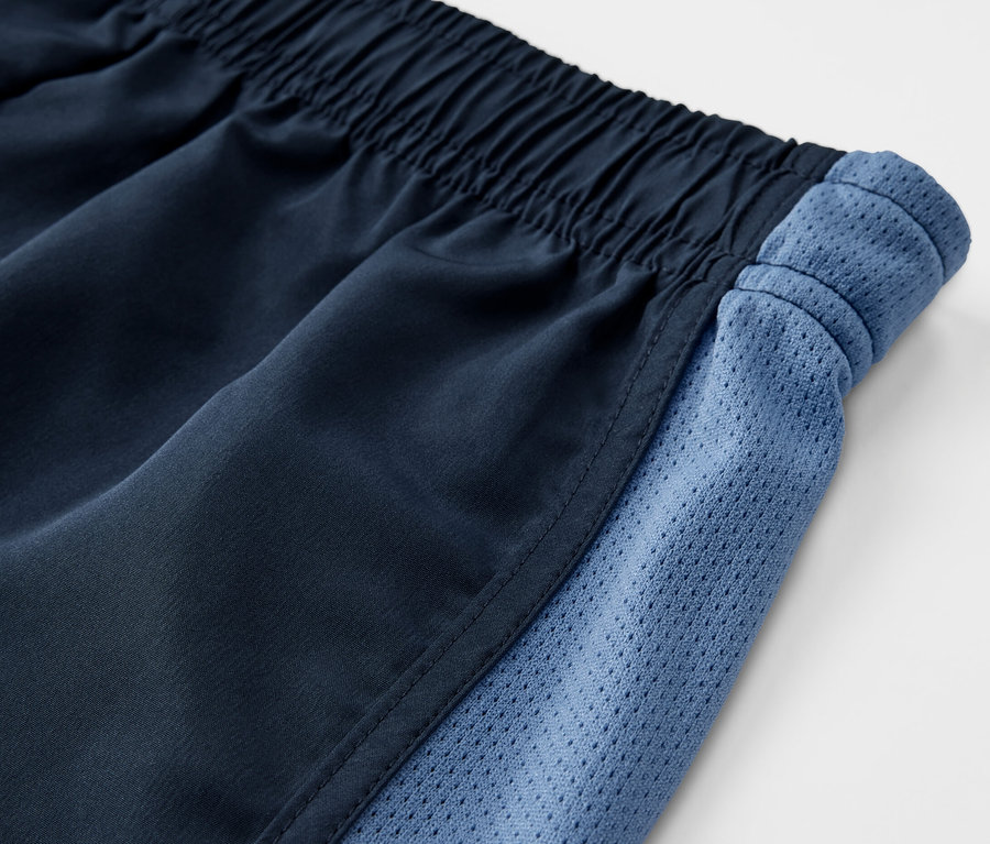 Gros plan sur un lot de 2 shorts de sport pour enfants avec une taille élastique et des empiècements latéraux en mesh bleu.