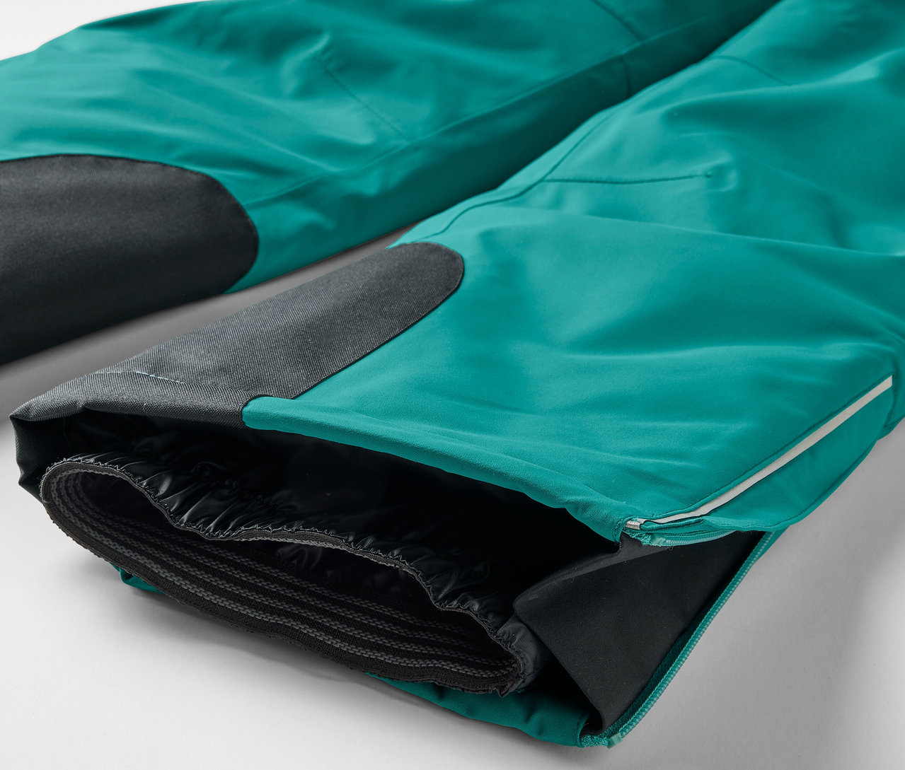 Gros plan sur un pantalon turquoise avec des patchs noirs aux genoux et un ourlet de jambe ouvert révélant une doublure noire.