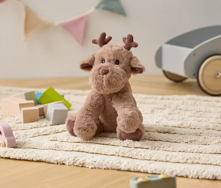 Un élan en peluche est assis sur un tapis crème parmi des jouets en bois et un chariot gris.