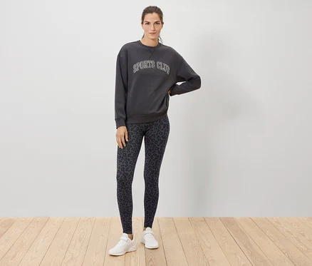 Une femme se tient sur un plancher en bois et porte un sweat-shirt gris avec l'inscription SPORTS CLUB, un legging à motif animalier et des baskets blanches.