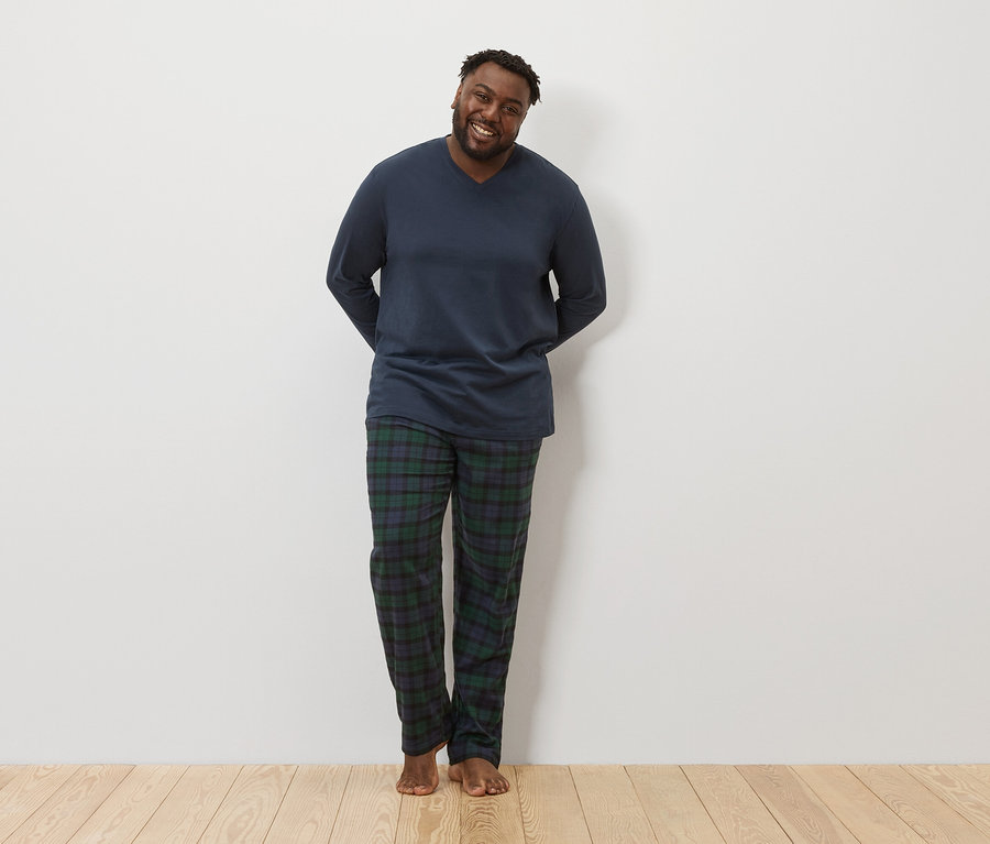 Un homme à la peau foncée se tient debout et sourit à la caméra. Il porte un haut de pyjama bleu marine à manches longues et un pantalon de pyjama à carreaux.