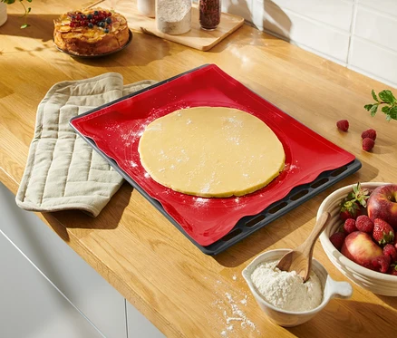 Sur un comptoir de cuisine se trouvent un tapis de cuisson réutilisable rouge avec de la pâte, une manique, un saladier de fruits et un gâteau.