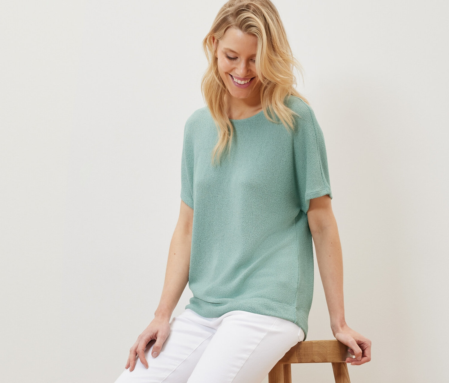 Une femme blonde est assise sur un tabouret en bois, portant un t-shirt structuré sauge et un pantalon blanc.