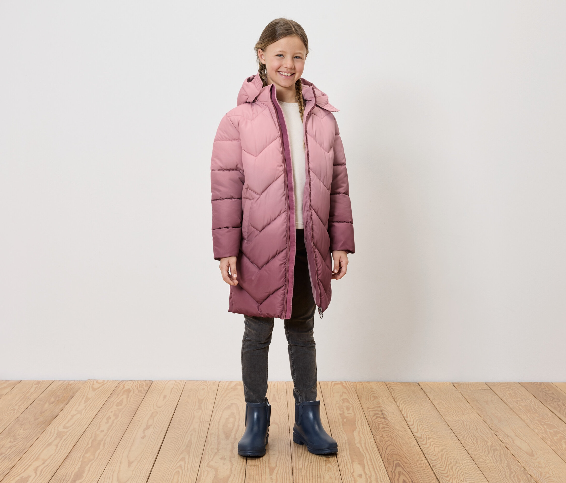 Une fille portant un long manteau à capuche matelassé, un jean gris et des bottes de pluie bleues, se tenant sur un plancher en bois devant un mur blanc.