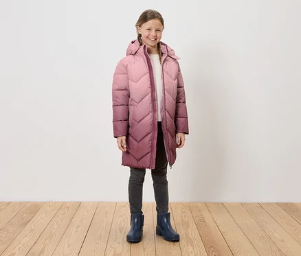 Une fille portant un long manteau à capuche matelassé, un jean gris et des bottes de pluie bleues, se tenant sur un plancher en bois devant un mur blanc.