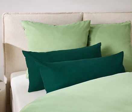 Deux oreillers verts et deux coussins oblongs vert foncé sont posés sur un lit avec une tête de lit crème et des draps vert clair.