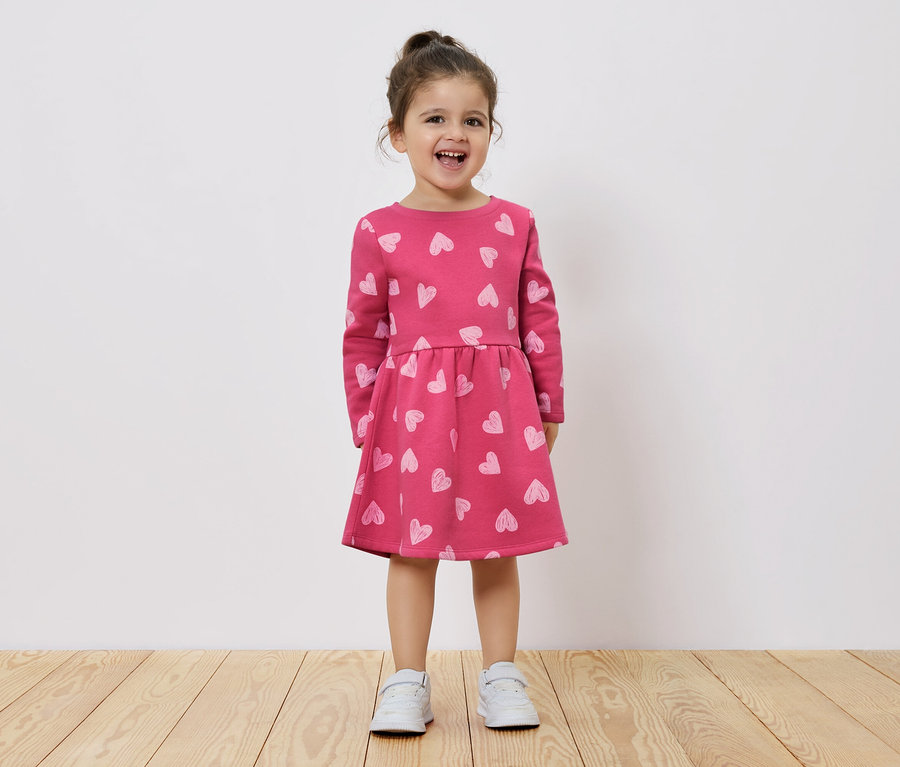 Une petite fille souriante se tient sur un plancher en bois et porte une robe rose à manches longues avec un imprimé de coeurs.