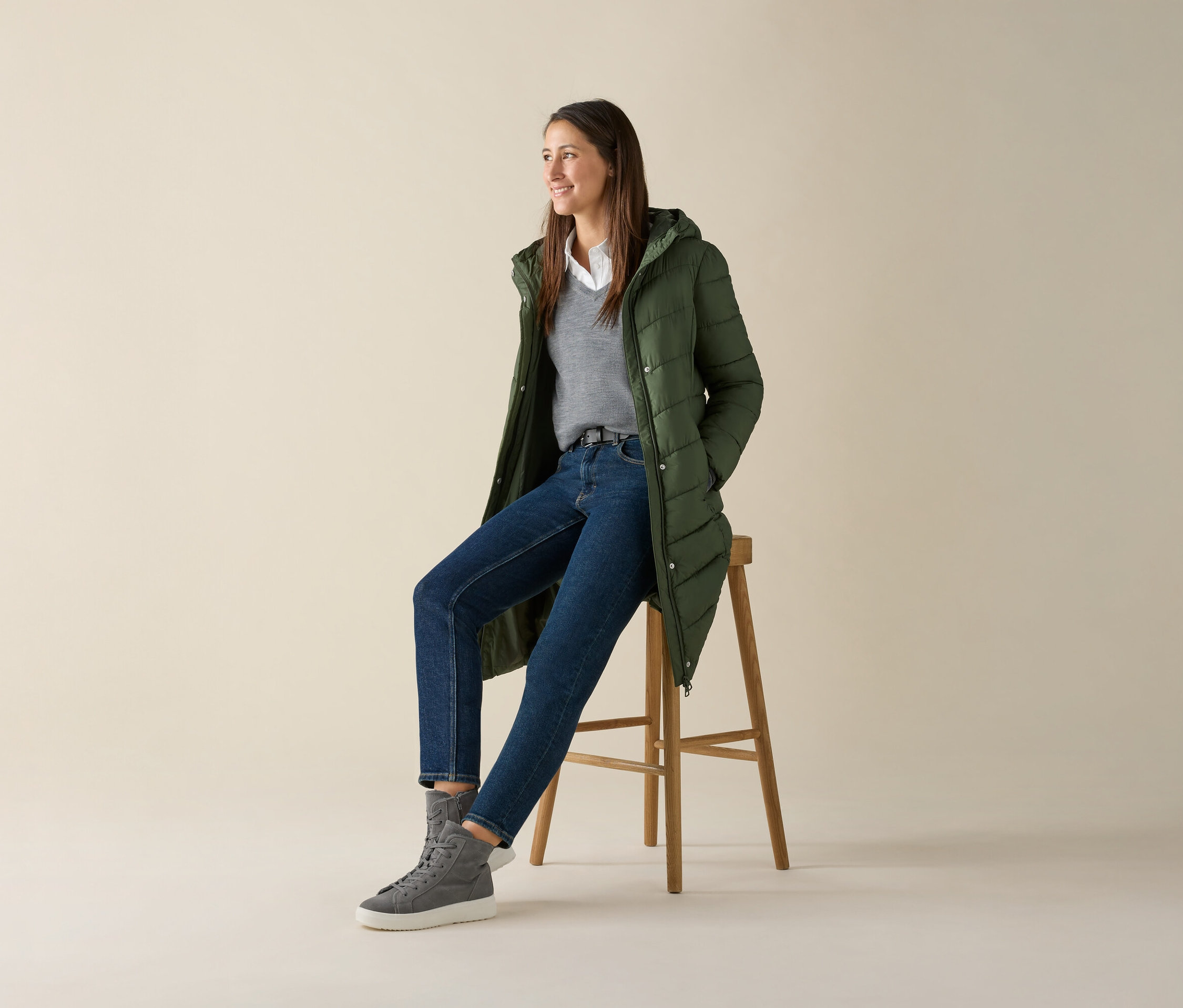 Une femme est assise sur un tabouret et porte un pull gris en maille fine de laine mérinos, un jean et une veste verte.