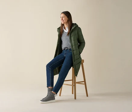 Une femme est assise sur un tabouret et porte un pull gris en maille fine de laine mérinos, un jean et une veste verte.