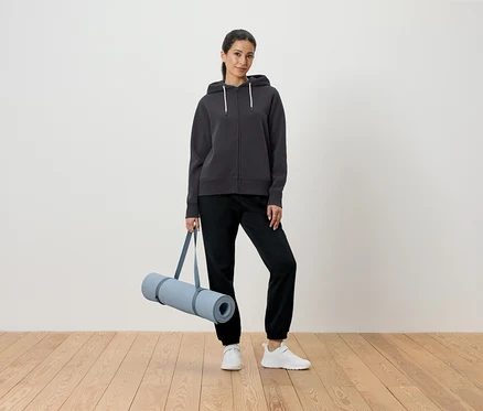 Femme tenant un tapis de yoga dans un sweat à capuche gris et un pantalon noir.