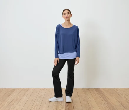 Une femme se tient sur un plancher en bois, portant un haut bleu et un pantalon noir.